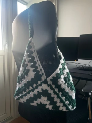 Grön och vit virkad axelväska - Unik handgjord axelväska i grönt och vitt med coolt virkat mönster. Väskan har bred axelrem och rymlig form, perfekt för att bära allt du behöver. Materialet är mjukt garn som ger en mysig och trendig look. Perfekt accessoar för dig som vill sticka ut.
