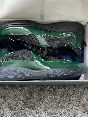 Gröna sneakers från Prada - Snygga sneakers från Prada i blankt grönt och svart material med meshpaneler och svarta snören. Skorna har en chunky sula i grått och svart och en modern, sportig design som sticker ut. Perfekta för dig som vill ha något unikt och trendigt i garderoben.