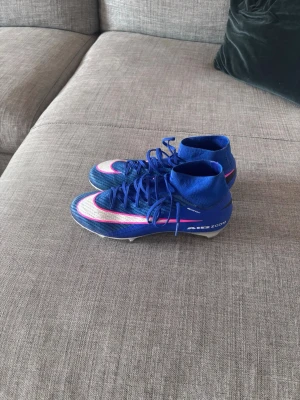 Nike blåa Mercurial fotbollsskor - Använda 1 gång endast på gräss Säljer ett par Nike Mercurial fotbollsskor i blått med vita och rosa detaljer. Skorna har hög ankelkrage, snörning och texten 'AIR ZOOM' på sidan. Perfekta för dig som vill ha grym kontroll och stil på planen.