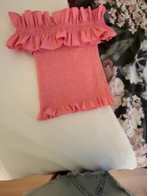 Rosa offshoulder blus med volanger - Supersöt rosa offshoulder topp med smockad kropp och bred volang upptill. Toppen är figurnära och elastisk, vilket ger en tight och bekväm passform. Materialet känns mjukt och stretchigt, perfekt för sommarens alla tillfällen.