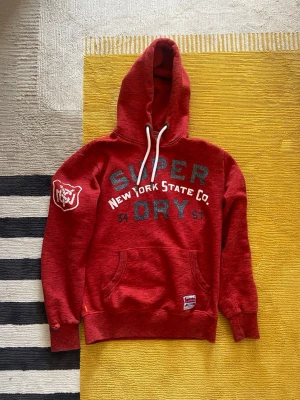 Superdry hoodie - Röd hoodie från Superdry med tryck i blått och vitt framtill, samt broderad björn på ärmen. Klassisk känguruficka, vita dragsnören och ribbade muddar. Perfekt för en avslappnad streetstyle-look.
