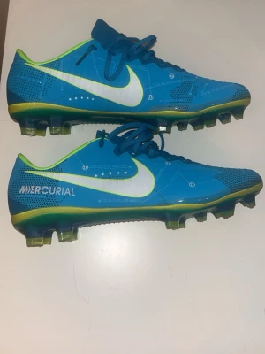 Nike Mercurial blå fotbollsskor - Säljer ett par Nike Mercurial fotbollsskor i blått med vita och neongröna detaljer. Skorna har låg profil, snörning och mönstrad sula för bra grepp på planen. Swoosh-logga på sidan och Mercurial-tryck baktill. Perfekta för dig som vill sticka ut på fotbollsplanen.