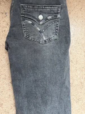 Grå jeans med broderad bakficka lågmidjade  - Snygga grå jeans med unikt broderi på bakfickan och silverfärgad knapp. Klassisk femficksmodell med dragkedjegylf och bälteshällor. Jeansen har en tvättad look. Från Gina lågmidjade 