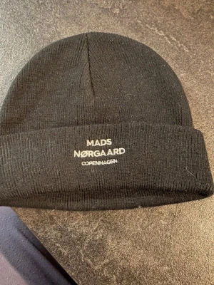 Svart mössa från Mads Nørgaard bra skick one size  - Snygg svart mössa från Mads Nørgaard med broderad logga framtill. Klassisk ribbstickad design och uppvikt kant. Perfekt accessoar för att hålla värmen och addera stil till din outfit.