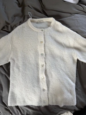 Vit ribbstickad kofta från Gina Tricot - Mysig vit ribbstickad kofta från Gina Tricot med rund halsringning och pärlemorknappar framtill. Koftan har lång ärm och är tillverkad i ett mjukt, fluffigt material som ger en avslappnad och trendig look. Storlek L. Knappt använd