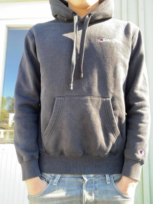 Svart hoodie från Champion - Svart hoodie från Champion med broderad logga på bröstet och ärmen. Klassisk känguruficka framtill, justerbar huva med dragsko och ribbade muddar. Perfekt för en avslappnad streetwear-look.