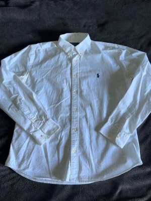 Vit skjorta från Polo Ralph Lauren - Klassisk vit skjorta från Polo Ralph Lauren i slim fit-modell. Skjortan har lång ärm, knappar framtill och broderad logga på bröstet. Tillverkad i bomull och passar perfekt till en clean och stilren look.