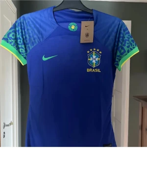 Blå Brasilien fotbollströja Nike - Snygg blå fotbollströja från Nike med Brasilien-logga och stjärnor på bröstet. Ärmarna har mönster i blått och grönt med gula detaljer. Perfekt för dig som älskar fotboll och vill stötta Brasilien. Materialet är lätt och andas bra.