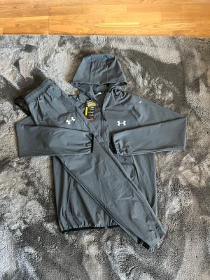 Under armour tracksuit  - Snyggt grått träningsset från Under Armour med matchande byxor och jacka med huva. Setet har dragkedja framtill, elastiska muddar och reflekterande detaljer på både jacka och byxor. Perfekt för löpning eller gym. Materialet är lätt och snabbtorkande. Skriv gärna vid frågor :)