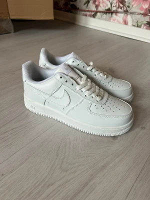 Nike Air Force 1 vita sneakers - Klassiska Nike Air Force 1 sneakers i helvitt läder med perforerad tå, vit sula och ikonisk Swoosh på sidorna. Skorna har lågt skaft och snörning framtill. Perfekta för en clean och stilren look.