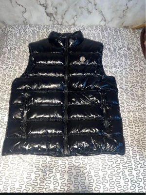 Svart glansig dunväst från Moncler - Snygg svart glansig dunväst från Moncler med hög krage och dragkedja framtill. Västen har två fickor med dragkedja och Moncler-logga på bröstet. Perfekt för lager på lager och ger en trendig look.