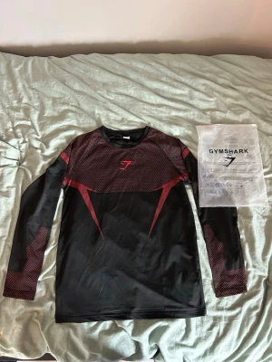 Svart och röd långärmad Gymshark tröja - Sportig långärmad tröja från Gymshark i svart med röda detaljer och mönster. Tillverkad i ett stretchigt syntetmaterial som andas, perfekt för träning. Gymshark-logga på bröstet och cool design på ärmar och axlar.