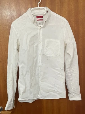 Vit skjorta från Hugo Boss Extra Slim - Klassisk vit skjorta från Hugo Boss med diskreta färgstänk i tyget. Modellen är Extra Slim Fit och har en bröstficka, knappar framtill och lång ärm. Perfekt för dig som gillar en clean och modern look.
