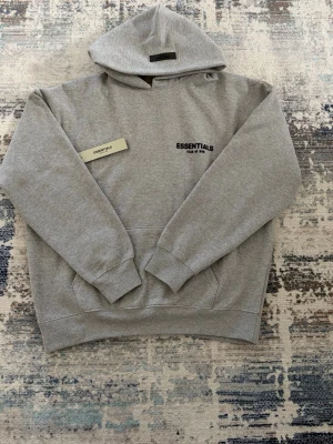 Grå Essentialsw Fear of God hoodie - Snygg grå hoodie från Essentials Fear of God med svart logga på bröstet. Klassisk passform med huva och känguruficka framtill. Tillverkad i mjukt bomullsmaterial som är perfekt för en avslappnad streetwear-look.