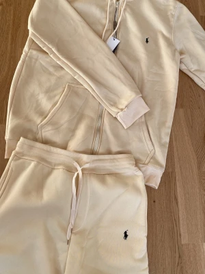 Beige hoodie och byxor från Polo Ralph Lauren - Beige hoodie från Polo Ralph Lauren med dragkedja, känguruficka och broderad svart logga på bröstet. Tillverkad i mjukt material med ribbade muddar och klassisk huva. Perfekt för en chill och stilren look. Byxor tillkommer. Hör av dig vid minsta fråga 💖💖
