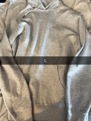 Ljusgrå basic hoodie med huva - En stilren ljusgrå hoodie med huva och dragsko. Hoodien har en klassisk passform, ribbade muddar vid ärmslut och nederkant samt en stor magficka framtill. Perfekt för en avslappnad och enkel look.
