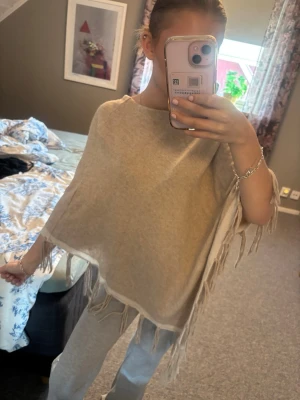 Beige poncho med fransar - !!Vet tyvärr inte storleken men den sitter ungefär som en S skulle jag säga!! Snygg beige poncho med fransar längs kanterna och ljusare kantdetaljer. Perfekt att slänga över en tröja för en avslappnad look. Mjuk och skön, troligtvis i stickat material.
