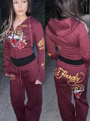 Ed Hardy tracksuit, size XS - Säljer ett vinrött loungewear-set från Ed Hardy med ikoniskt tigertryck och rosor på tröjan samt guldiga loggor på både byxa och hoodie. Setet består av en zip-hoodie med dragsko och matchande byxor med resår i midjan. Materialet är mjukt och skönt, perfekt för chill dagar.