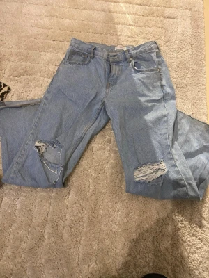 Ljusa slitna jeans från MNG Denim - Säljer ett par ljusblå jeans från MNG Denim med raka ben och slitningar på båda knäna. Klassisk femficksmodell i bomullsdenim med snygga detaljer och avslappnad passform. Perfekta för en trendig och avslappnad stil.