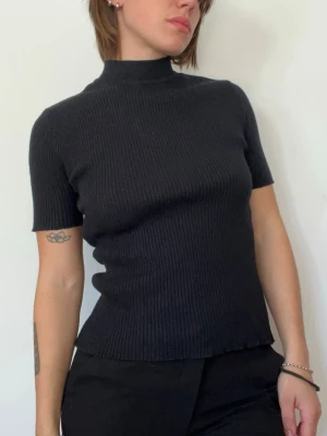Svart ribbstickad topp med kort ärm - Säljer en svart ribbstickad topp med korta ärmar och hög hals. Tröjan har en figurnära passform och är tillverkad i ett mjukt, stretchigt material. Perfekt för en stilren och enkel look. Det står storlek L men den är väldigt stretchig så skulle säga att den passar M och S också 
