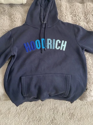 Mörkblå HOODRICH hoodie med brodyr - Snygg mörkblå hoodie från HOODRICH med broderad logga i blå och turkosa nyanser över bröstet. Klassisk känguruficka framtill och justerbar huva med dragsko. Perfekt för en chill och avslappnad stil.