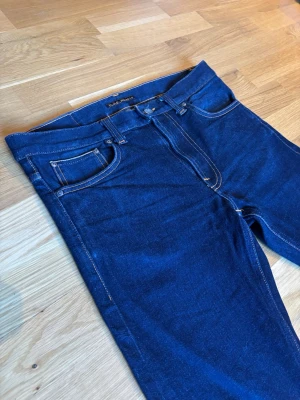 Nudie Jeans Lean Dean – Selvedge (Slim Tapered) - Klassiska Lean Dean i premium Selvedge-denim. En slim passform som möter hantverket hos kantstickad denim. Jeansen har den klassiska orangea selvedge-tråden på insidan av benet.  Modell: Lean Dean  Material: Comfort Selvedge (stretch för komfort men med selvedge-look)  Passform: Slim Tapered W31 L30  Skick: Mycket gott skick, väl omhändertagna.
