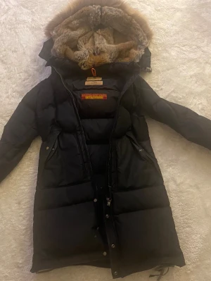 Svart lång dunjacka Parajumpers - Svart lång dunjacka från Parajumpers med pälsdetalj på huvan, dragkedja och tryckknappar framtill. Jackan är varm, mycket fin i modellen och i bra skick.