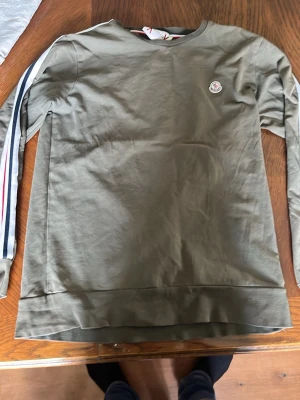Olivgrön sweatshirt från Moncler - Olivgrön sweatshirt från Moncler med rund hals och broderad Moncler-logga på bröstet. Ärmarna har ränder i vitt, blått och rött längs sidorna. Tillverkad i mjukt bomullsmaterial och har en stilren, avslappnad passform.