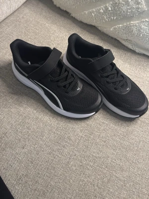 Svarta puma gympa skor - Svarta och vita barnsneakers från Puma med bekväm passform, kardborreband och lätt dämpad sula. Perfekta för lek, skola och vardagsbruk.