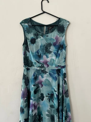 Blommig grön klänning med bälte - Ny snygg långklänning med blommigt mönster i blå, lila och gröna toner. Klänningen är ärmlös, har rund halsringning och ett smalt bälte i midjan. Perfekt för dig som gillar färg och mönster! Nypris 2899