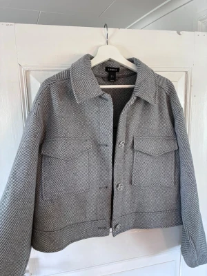 Grå mönstrad cardiganjacka Lindex - Snygg grå overshirt från Lindex i storlek L. Jackan har ett klassiskt fiskbensmönster, stora bröstfickor med lock och knäppning framtill. Rak passform och bred krage ger en trendig look. Perfekt att slänga över en hoodie eller t-shirt.