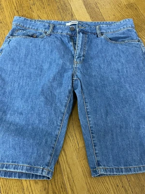 Blå denimshorts från Asos - Klassiska blå denimshorts från Asos med fem fickor, bälteshällor och knappgylf. Modellen har en rak passform och går ner till knäna. Perfekta för varma dagar och enkel att matcha med olika stilar.