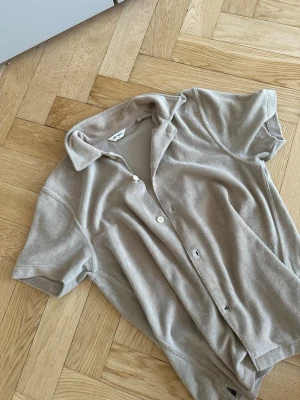 Beige kortärmad skjorta i frotté - Snygg beige skjorta från East west i mjuk frotté med korta ärmar och klassisk krage. Perfekt för en avslappnad och trendig look. Skjortan har knappar framtill och en loose passform. Tröjan e Xs i vuxen, jag är 173 och den passar mig perfekt. Tröjan har lite solblekta märken i insidan kragen men de syns inte vid användning
