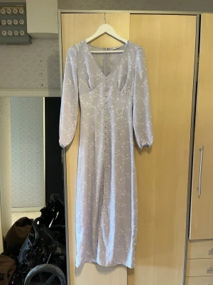 Ljuslila långklänning med blommor - Söt långklänning i ljuslila med vitt blommönster och v-ringning. Klänningen har långa ärmar och smal midja för en feminin siluett. Även en slits framtill. Perfekt för vår och sommar, tillverkad i ett mjukt och lätt material. 💜