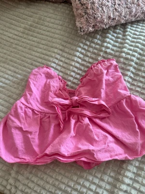 Rosa bandeau topp med rosett - Söt rosa bandeau topp med volangkant och en stor rosett framtill. Toppen är ärmlös och har en luftig passform, perfekt för varma dagar. 