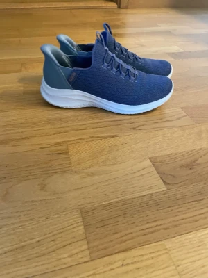 Blå Skechers sneakers med vit sula - Snygga blå sneakers från Skechers med stickad ovandel och elastiska snören. Skorna har en vit, lättviktig sula och ljusblå detaljer vid häl och insida. Perfekta för en sportig och avslappnad stil.