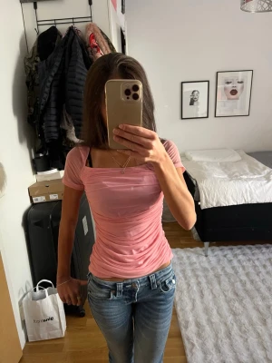 Ljusrosa tight topp med kort ärm - Säljer en ljusrosa tight topp med korta ärmar och rynkade sidor. Toppen har en feminin, figurnära passform och är tillverkad i ett stretchigt syntetmaterial. Toppen är köpt i Miami och lappen har dragits av, så vet dessvärre inte vart den är köpt!