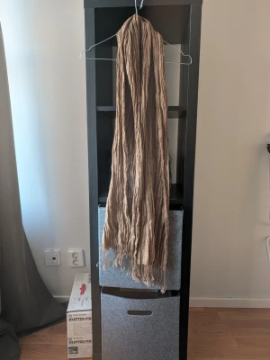 Beige crinklad halsduk DKNY - Lång och luftig halsduk i beige med crinklad struktur och fransar i ändarna. Perfekt för att ge en avslappnad och trendig touch till din outfit. Materialet känns lätt och passar bra till lager-på-lager-stil.