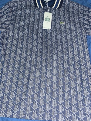 Blå mönstrad pikéskjorta från Lacoste - Snygg blå pikéskjorta från Lacoste med vitt geometriskt mönster och klassisk krage med ränder. Skjortan har korta ärmar, knappar vid halsen och den ikoniska krokodilloggan på bröstet. Perfekt för dig som gillar sportig och trendig stil. Helt ny med tags och påse medföljer. Den är aldrig använd köpte fel storlek 