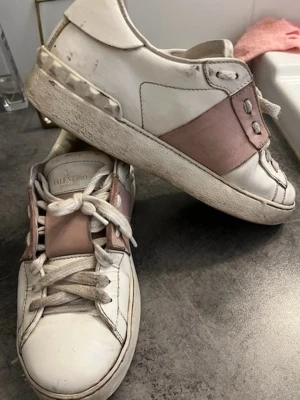 Valentino vita sneakers med rosa detalj - Snygga vita sneakers från Valentino med bred rosa läderdetalj över snörningen. Klassisk låg modell med rund tå och vita skosnören. Skorna har en chunky sula och diskreta sömmar som ger en stilren look.