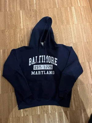 Mörkblå hoodie med Baltimore-tryck - Snygg mörkblå hoodie med vit text där det står 'Baltimore Maryland EST. 1729' på bröstet. Klassisk modell med huva och dragsnören samt en stor magficka. Perfekt för en avslappnad och cool stil.