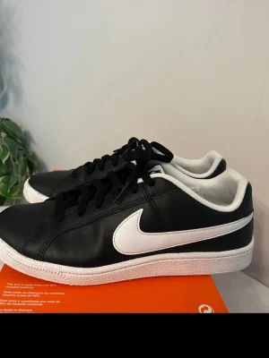 Nike sneakers court low - Snygga svarta sneakers från Nike med klassisk vit swoosh på sidan och vit sula. Skorna har låga skaft, svarta skosnören och en stilren design. Ovandelen är i slätt material och insidan är vadderad för extra komfort. Perfekta för en sportig och clean look. STORLEK 41 men små i storlek