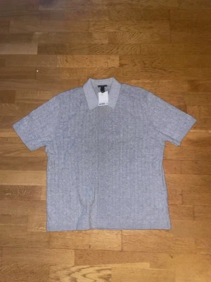 Grå kabelstickad skjorta från H&M - Snygg grå kabelstickad skjorta från H&M med korta ärmar och klassisk krage. Skjortan har en mjuk och bekväm känsla, tillverkad i bomullsmix. Perfekt för en avslappnad men stilren look.