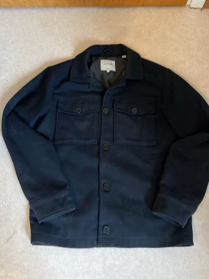 Mörkblå overshirt från Jack & Jones - Stilren mörkblå overshirt från Jack & Jones med klassisk krage och två stora bröstfickor med lock och knapp. Jackan har knäppning framtill och är tillverkad i ett mjukt, lite tjockare material som passar perfekt till lager-på-lager.