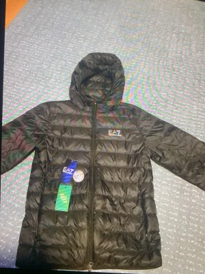 Svart dunjacka från EA7 Emporio Armani - Snygg svart dunjacka från EA7 Emporio Armani med huva och dragkedja framtill. Jackan har ett quiltat mönster och logga på bröstet. Perfekt för vår&sommsr och ger en sportig look. Materialet är lätt och smidigt, vilket gör den enkel att bära.