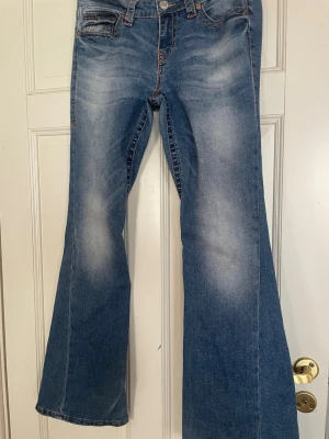 Blå bootcut jeans från True Religion - Snygga blå bootcut jeans från True Religion med klassisk femficksdesign och kontrastsömmar. Jeansen har utsvängda ben och dekorativa bakfickor med broderade detaljer. Perfekta för en avslappnad och trendig look.