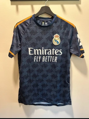 Real Madrid bortatröja Vini Jr. 7 - Snygg mörkblå Real Madrid bortatröja från Adidas med Vini Jr. och nummer 7 på ryggen. Tröjan har gula detaljer på axlar och ärmslut, klubbmärke på bröstet och Emirates Fly Better-tryck framtill. Perfekt för fotbollsfans!