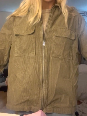Olivgrön overshirt i manchester - Säljer en olivgrön overshirt i manchestertyg med dragkedja framtill och två stora bröstfickor med lock. Overshirten har en avslappnad passform och klassisk krage, perfekt att slänga över en hoodie eller t-shirt. Inget synligt märke.
