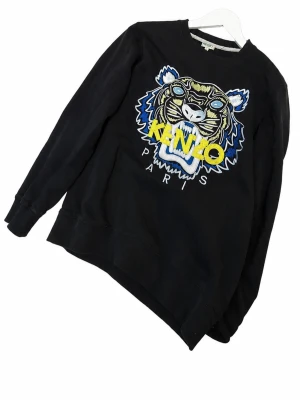 Svart Kenzo sweatshirt med tigertryck - Cool svart sweatshirt från Kenzo med stort broderat tigerhuvud och färgglada detaljer i blått, gult och vitt. Klassisk rund halsringning och ribbade muddar. Perfekt statement-plagg för dig som gillar streetwear och vill sticka ut.