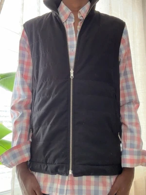Svart dunväst j.Lindeberg - Stilren svart dunväst med hög krage och silverfärgad dragkedja framtill,från J.Lindeberg. Västen har en klassisk rak passform och är perfekt att slänga över en skjorta eller hoodie. Materialet är quiltat och ger en snygg, puffig look.